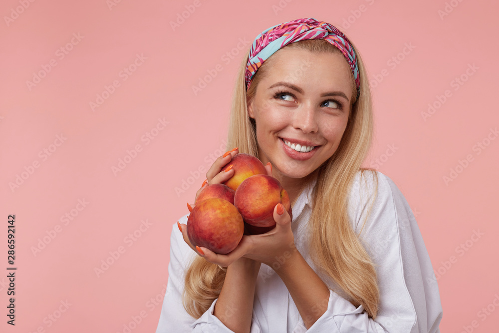 Attractive young woman in white shirt and colored headband with long blond hair looking aside joyfully, holding peaches in hands over pink background Attractive young woman in white shirt and colored headband with long blond hair looking aside joyfully, holding peaches in hands over pink background