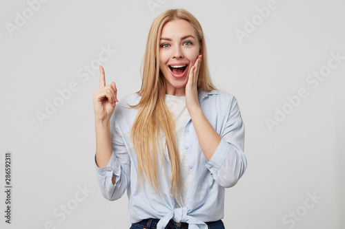 Close up portrait of young smiling woman in blue shirt keeping finger pointed upwards, making eureka gesture with index finger, isolated over white background