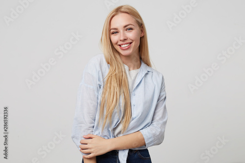 Indoor portrait of young attractive blonde long haired woman smiling to camera sincerely, posing over white background, wearing jeans and blue shirt