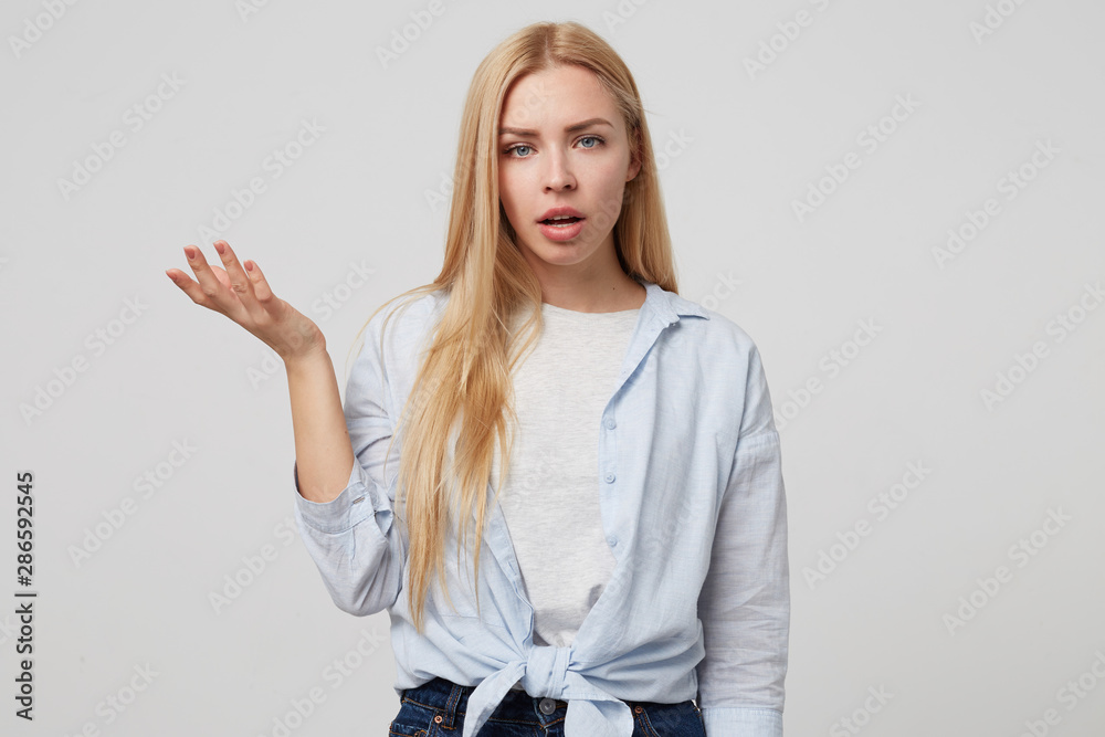 Horizontal indoor shot of angry young blonde female standing over white background with raised palm, looking annoyed and grumpy to camera Horizontal indoor shot of angry young blonde female standing over white background with raised palm, looking annoyed and grumpy to camera