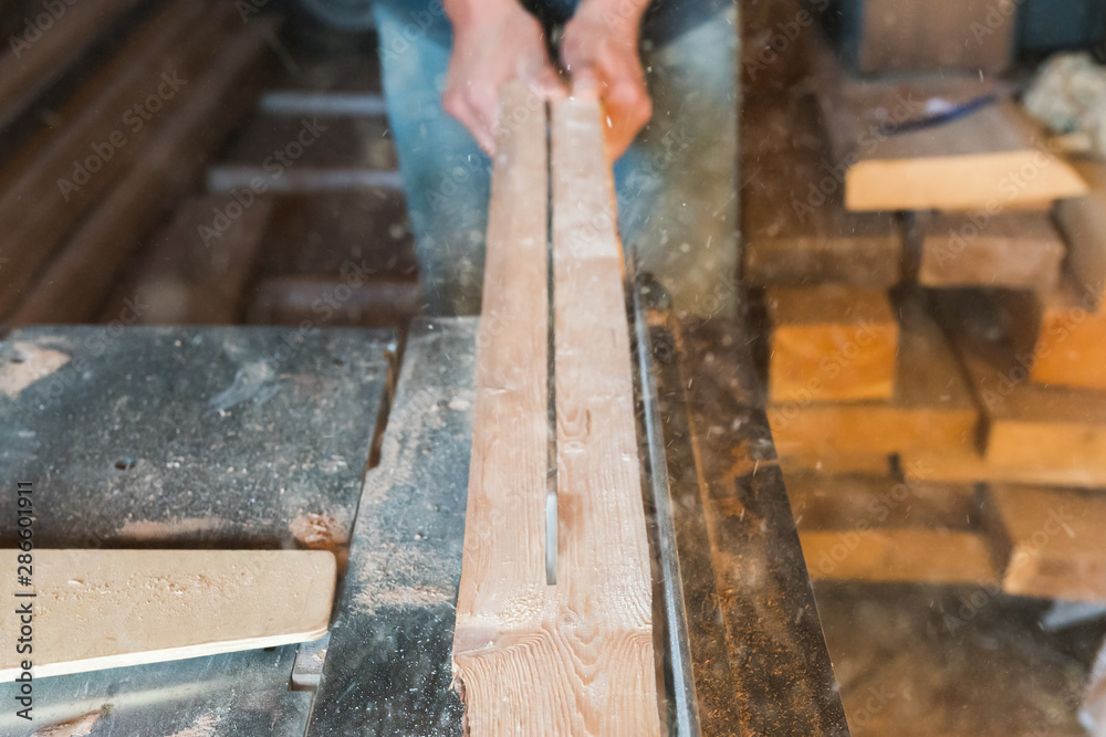 Woodworking machine process a wooden plank by a circular saw. Workers hands hold a wooden plank. selective focus. Motion blur. Manual wood processing concept. Woodworking machine process a wooden plank by a circular saw. Workers hands hold a wooden plank. selective focus. Motion blur. Manual wood processing concept.
