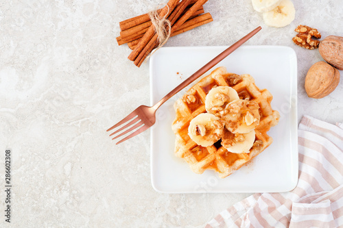 Breakfast waffle with bananas, walnuts and syrup. Above view table scene over a white marble background.