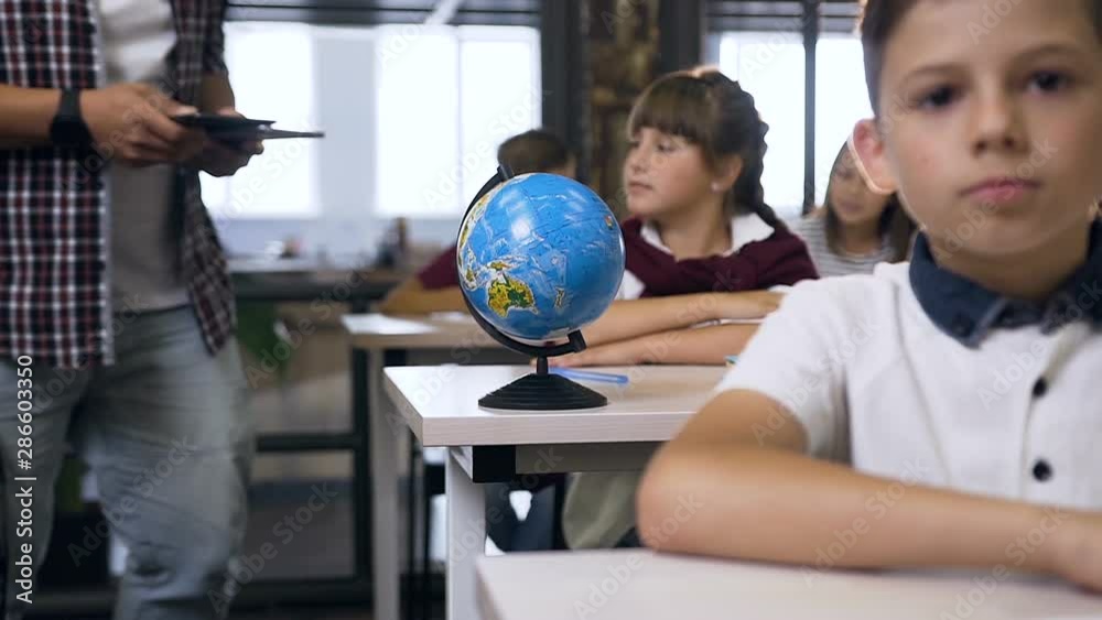 Male teacher of elementary school walking between desks puts computers tablet on desk pupils for learning Male teacher of elementary school walking between desks puts computers tablet on desk pupils for learning