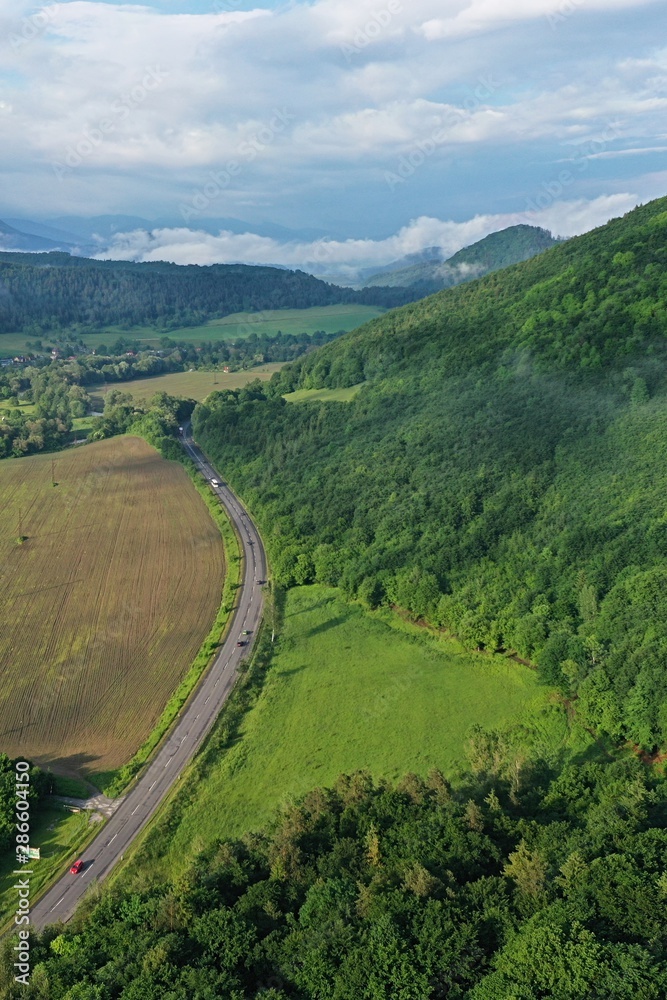 Aerial drone view of asphalt road between densely forested hill and plowed field, location near Slovenska Lupca, Banska Bystrica region, central Slovakia. Shot taken during cloudy summer day