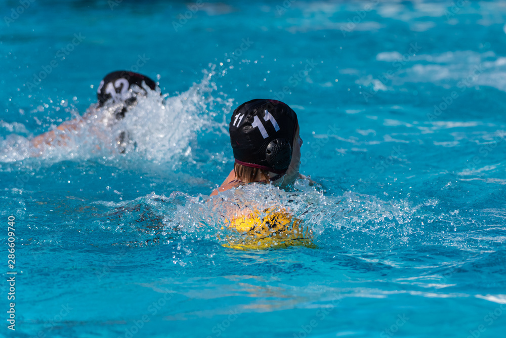 Water polo player in black uniform wearing number eleven swims with ball while looking back toward the defensive side.
