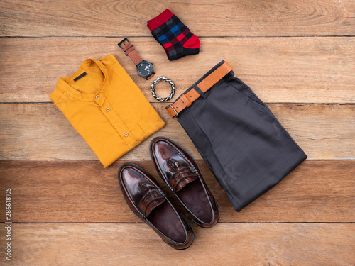 Men fashion casual clothing set on wooden background include brown loafer shoes, yellow shirt, belt ,watch, sunglasses, sock, bracelet and gray pants. Flat lay, top view.