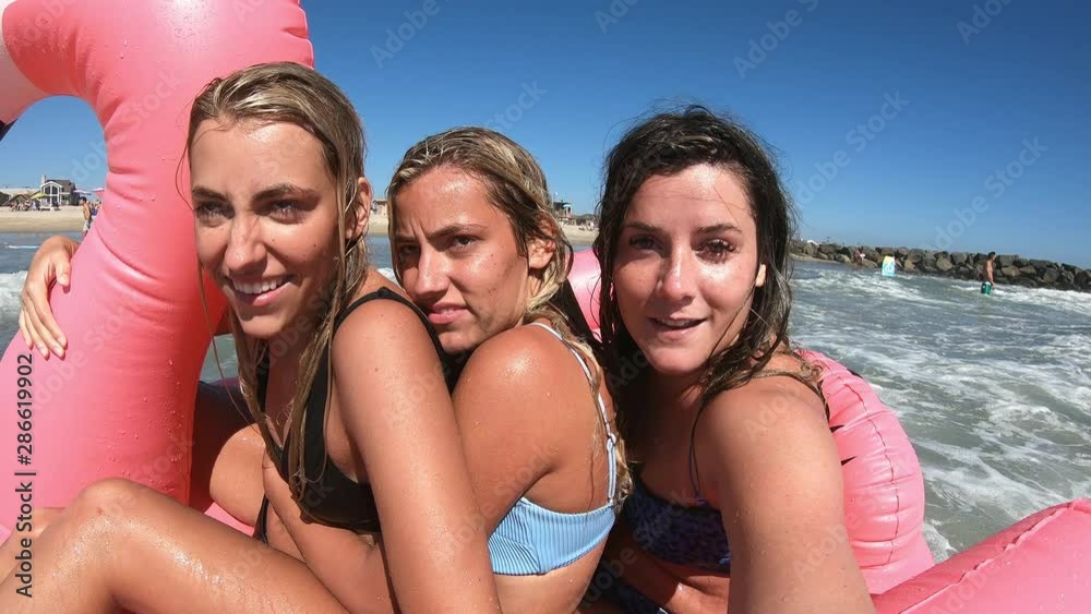Three young woman sitting on pink blowup flamingo floating in the ocean on hot summers day as they play in the waves.