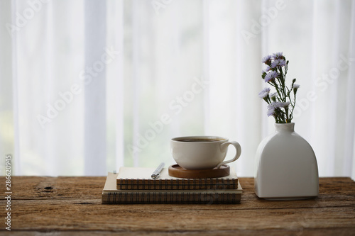 white coffee cup with diary notebook and flower in vase on wooden table on front of curtain