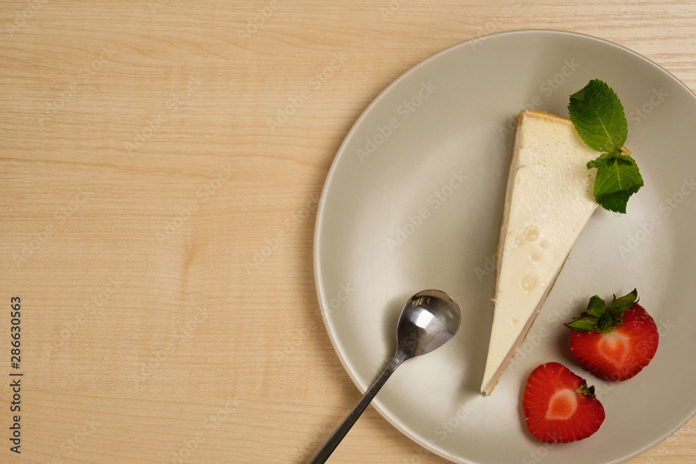 A plate with cheesecake, a spoon and strawberries stands on a light wooden table.