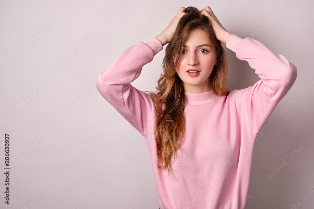 A girl in a pink sweater stands on a white background, looks at the camera, hands in her hair.