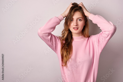 A girl in a pink sweater stands on a white background, looks at the camera, hands in her hair.