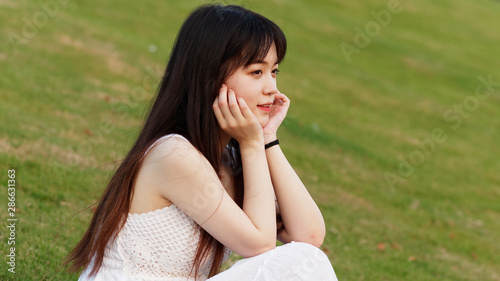 Portrait of beautiful Chinese young woman in white wedding dress sitting on grass field with hands on her face, beauty in summer.