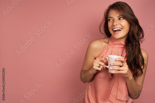 Tableau sur toile A dark-haired girl in a pink dress with a mug in her hand stands on a pink background and looks joyfully laughing sideways