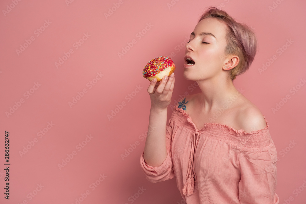 A girl with short pink hair in a blouse reaches for with a closed eyes and open mouth to a donut in her hand, on a pink background A girl with short pink hair in a blouse reaches for with a closed eyes and open mouth to a donut in her hand, on a pink background