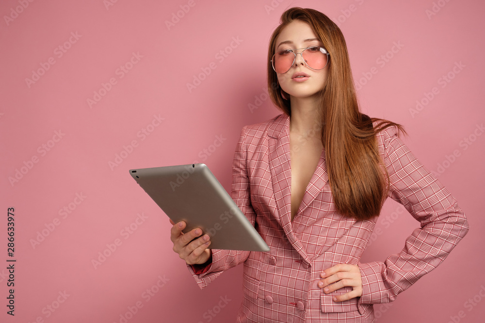 Fototapeta premium Red-haired girl in a jacket stands on a pink background with an electronic tablet and looks into the camera over glasses.