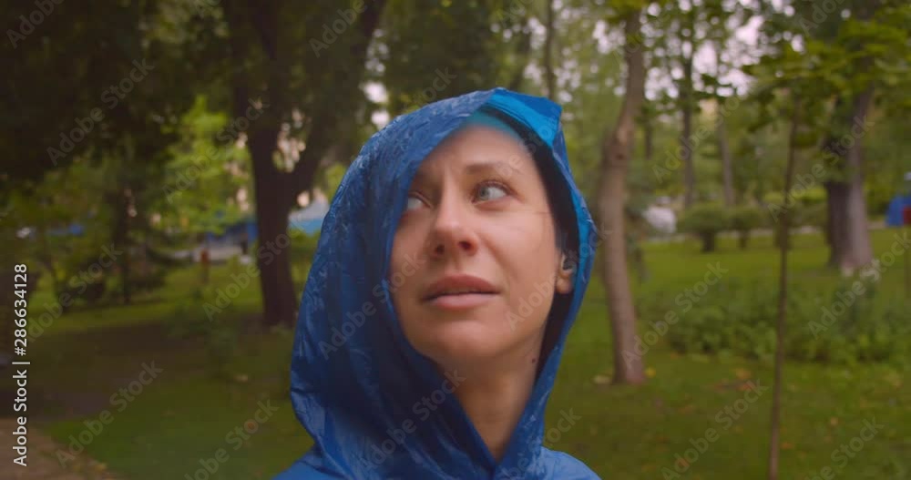 Closeup portrait of young caucasian female in blue raincoat looking at camera while it is raining standing in park outdoors Closeup portrait of young caucasian female in blue raincoat looking at camera while it is raining standing in park outdoors