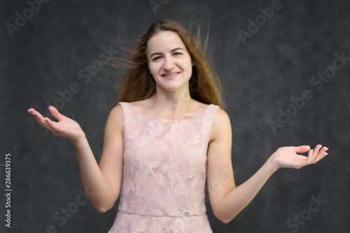 Studio portrait on the waist of a pretty student girl, a young brunette woman with long beautiful hair in a pink dress on a gray background. Smiling, talking, showing emotions