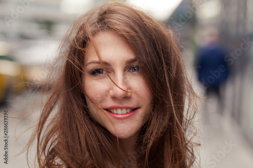Close portrait of elegant beautiful young smiling brunette woman with messy long hair scattered by the wind standing on the street