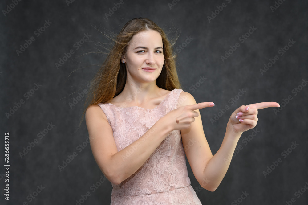 Fototapeta premium Studio portrait on the waist of a pretty student girl, a young brunette woman with long beautiful hair in a pink dress on a gray background. Smiling, talking, showing emotions