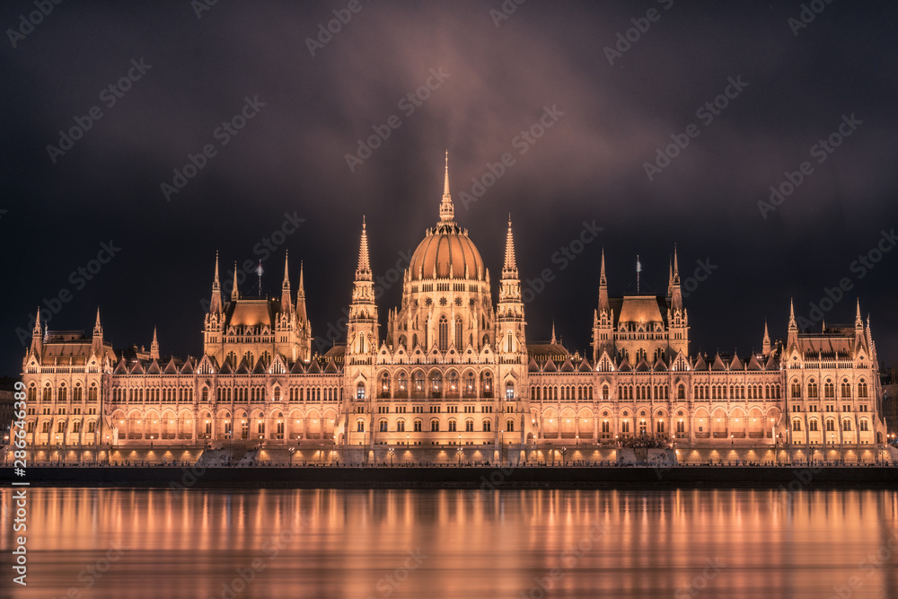 Illuminated Budapest parliament building at night with dark cloudy sky and reflection in Danube river