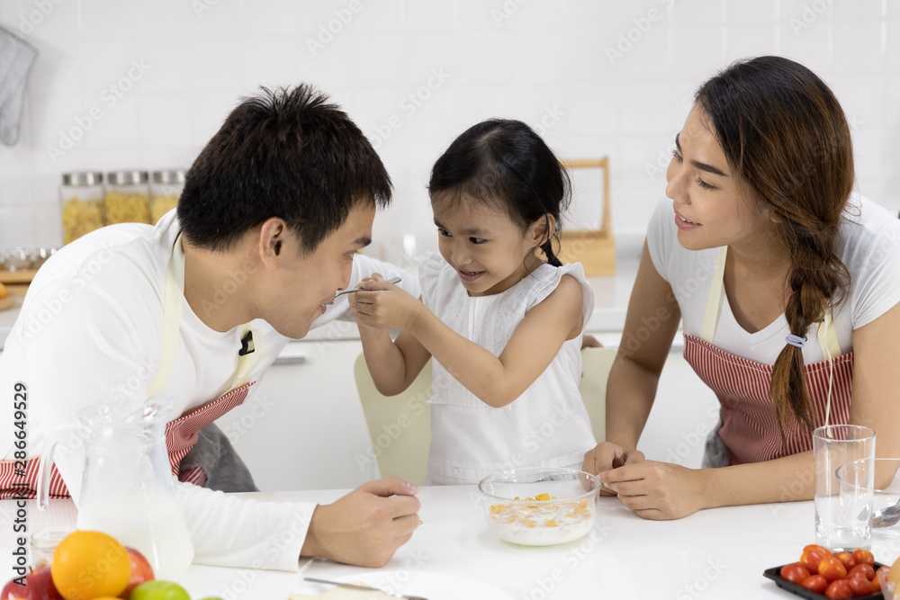 Happy Asian family make a cooking. Father, Mother and Daughter are eating breakfast, cereal with milk in the kitchen at home. Healthy food concept and happy holidays
