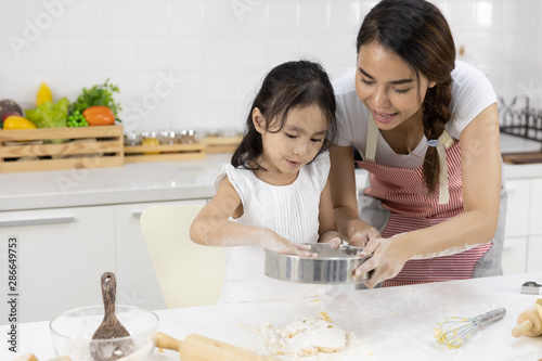 Happy Asian family Mother and Daughter are sieving flour preparing the dough, bake cookies in the kitchen in home. family cooking food Concept.