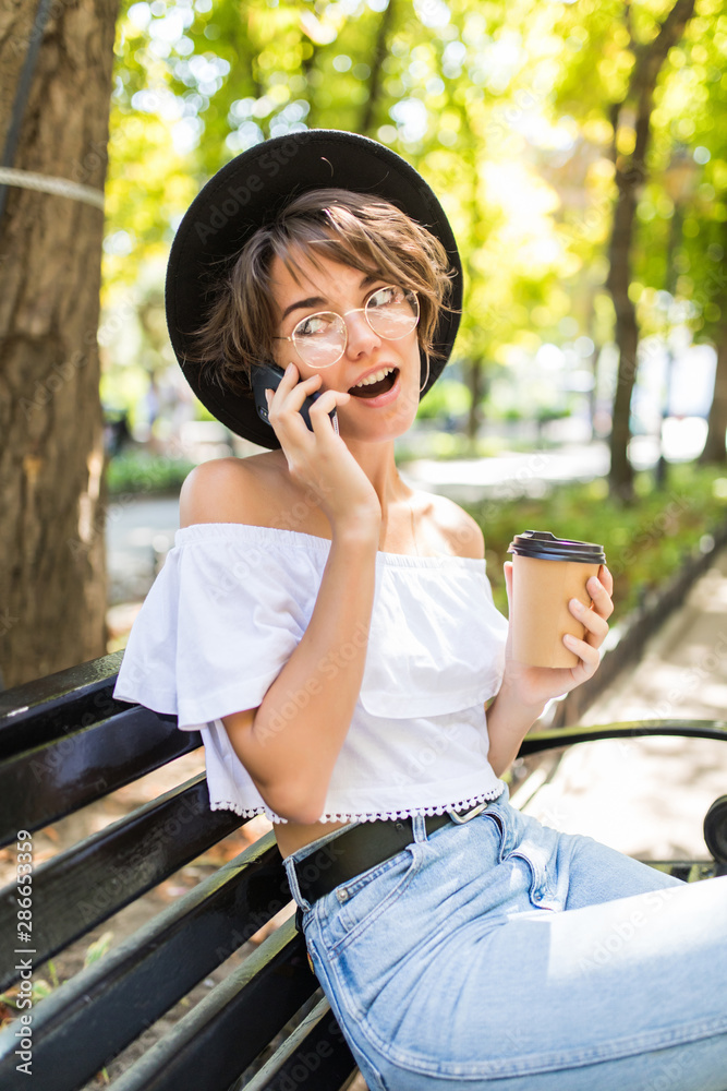 Smiling modern woman sitting on bench in green park on summer day and talking on smartphone with cup of takeaway coffee