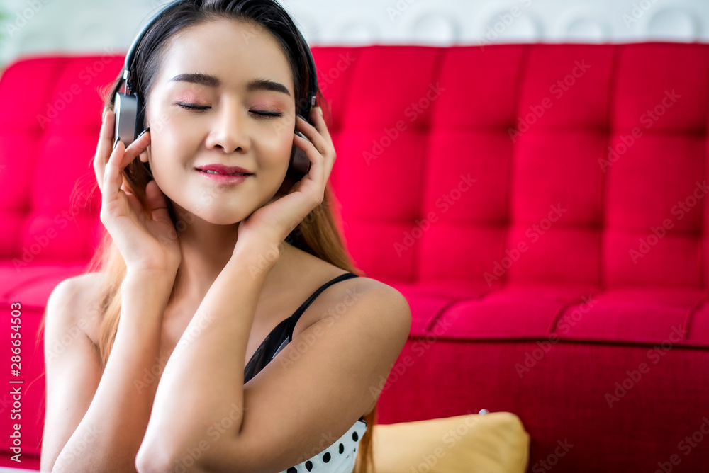 Young carefree attractive sensuality Asian woman enjoy to listening music connecting to laptop at living room with red sofa background.