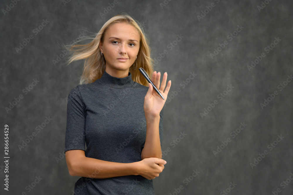 Fototapeta premium Studio portrait of a pretty blonde student girl, young woman in a gray sweater with a pen in her hands on a gray background. Talking, showing emotions.
