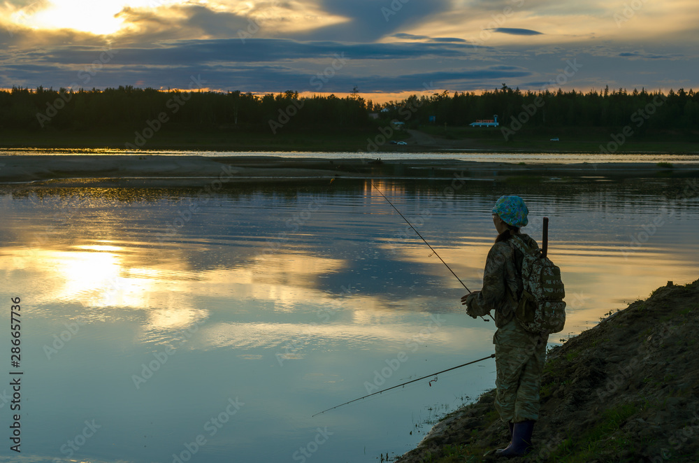 Girl fisherman tourist with a backpack holding one rod in his legs throws a second spinning on the background of a bright sunset in the wild taiga of Yakutia.