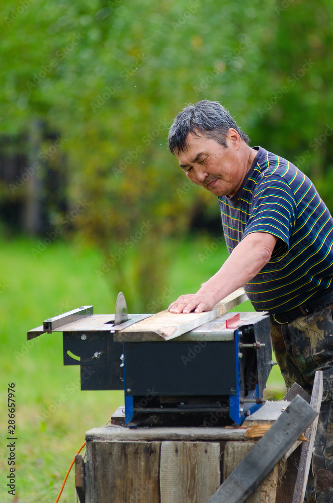 Yakut man neatly with a tense face sawing Board on a circular saw against a green garden in the Northern village.