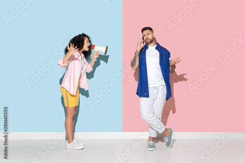Young emotional caucasian couple in bright casual clothes posing on pink and blue background. Concept of human emotions, facial expession, relations, ad. Woman screams with mouthpeace, man ignores her