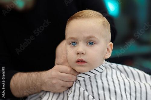 Cute blond caucasian baby boy with blue eyes in a barber shop after having haircut by hairdresser. Hands of stylist. Children fashion. Indoors, dark background, copy space.