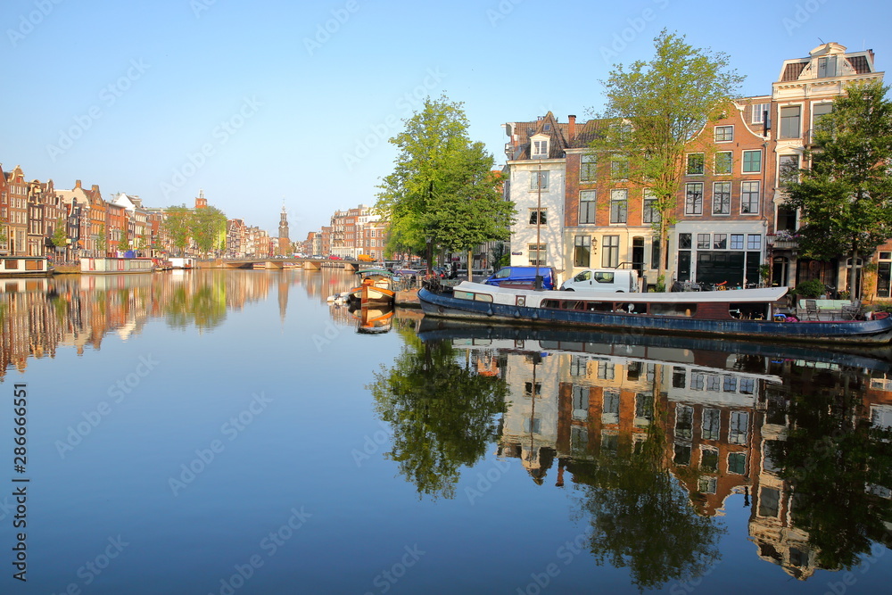 Colorful heritage buildings and houseboats, overlooking Zwanenburgwal canal and Amstel river with perfect reflections, with Munttoren (historic tower with a carillon) in the background, Amsterdam, Net