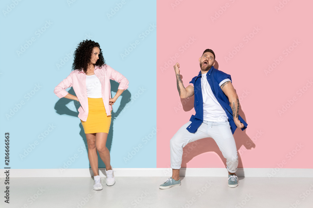 Young emotional caucasian couple in bright casual clothes posing on pink and blue background. Concept of human emotions, facial expession, relations, ad. Man's dancing and celebrating, woman is bored. Young emotional caucasian couple in bright casual clothes posing on pink and blue background. Concept of human emotions, facial expession, relations, ad. Man's dancing and celebrating, woman is bored.