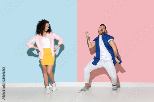 Young emotional caucasian couple in bright casual clothes posing on pink and blue background. Concept of human emotions, facial expession, relations, ad. Man's dancing and celebrating, woman is bored.