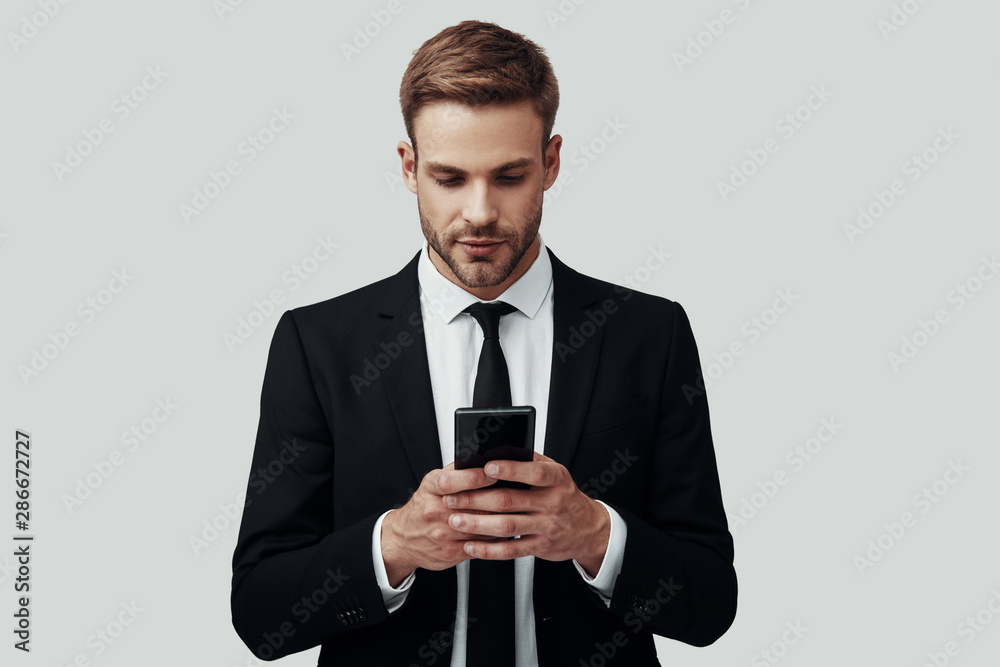 Handsome young man in formalwear using smart phone while standing against grey background Handsome young man in formalwear using smart phone while standing against grey background