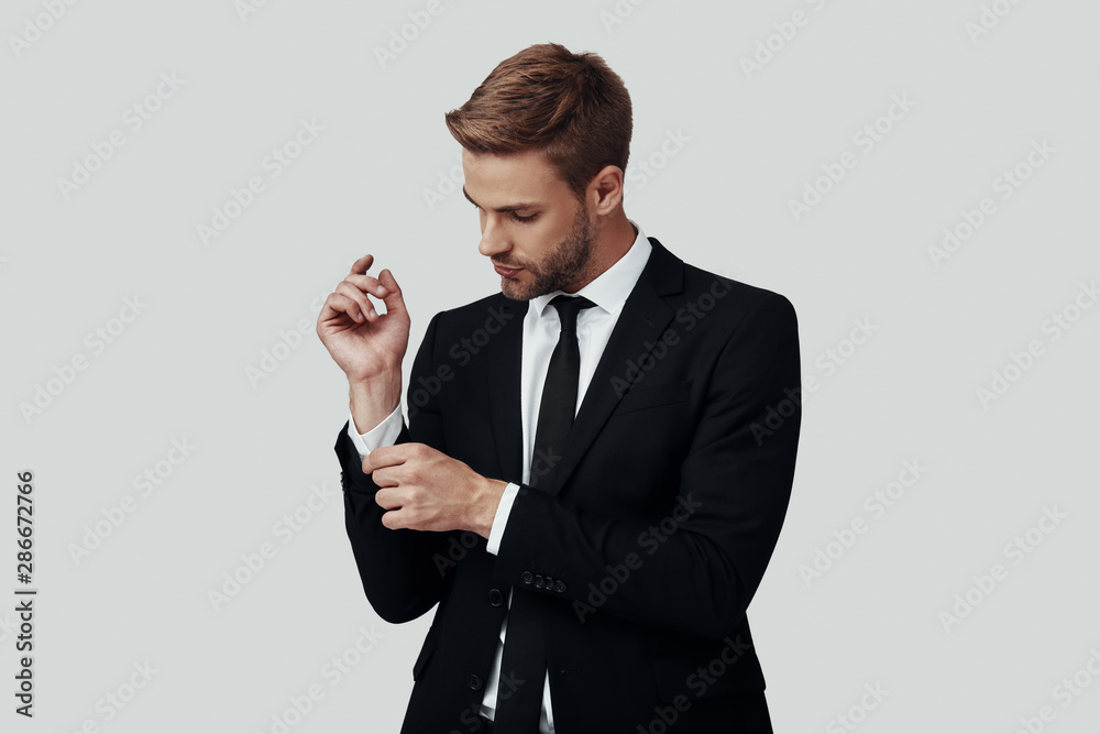 Handsome young man in formalwear adjusting sleeve while standing against grey background Handsome young man in formalwear adjusting sleeve while standing against grey background