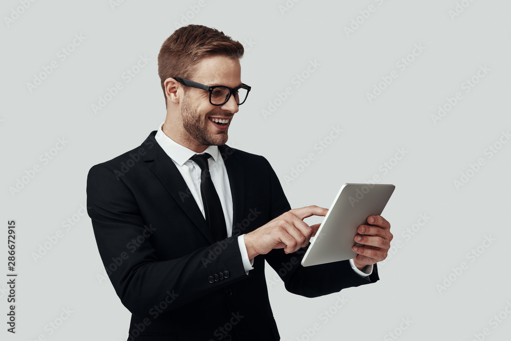 Handsome young man in formalwear working using digital tablet and smiling while standing against grey background