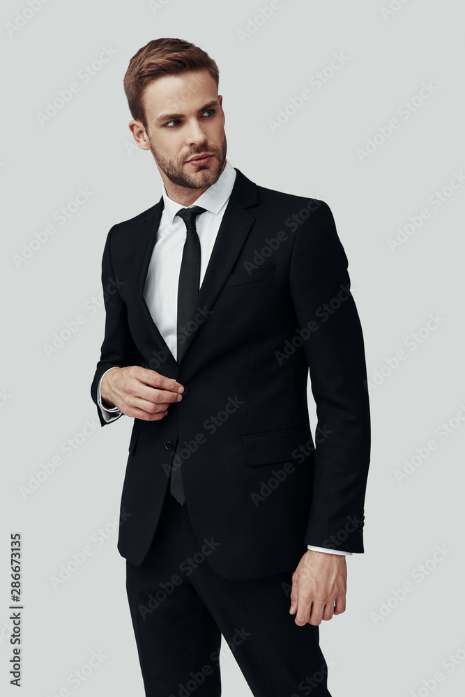 Handsome young man in formalwear looking away and adjusting jacket while standing against grey background Handsome young man in formalwear looking away and adjusting jacket while standing against grey background