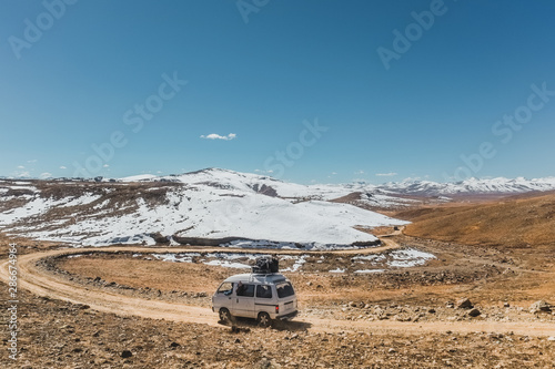 A local bus with luggage on the roof running along the unpaved road with landscape view of snow capped mountains at Deosai plain national park in Astore. Gilgit Baltistan, Pakistan.