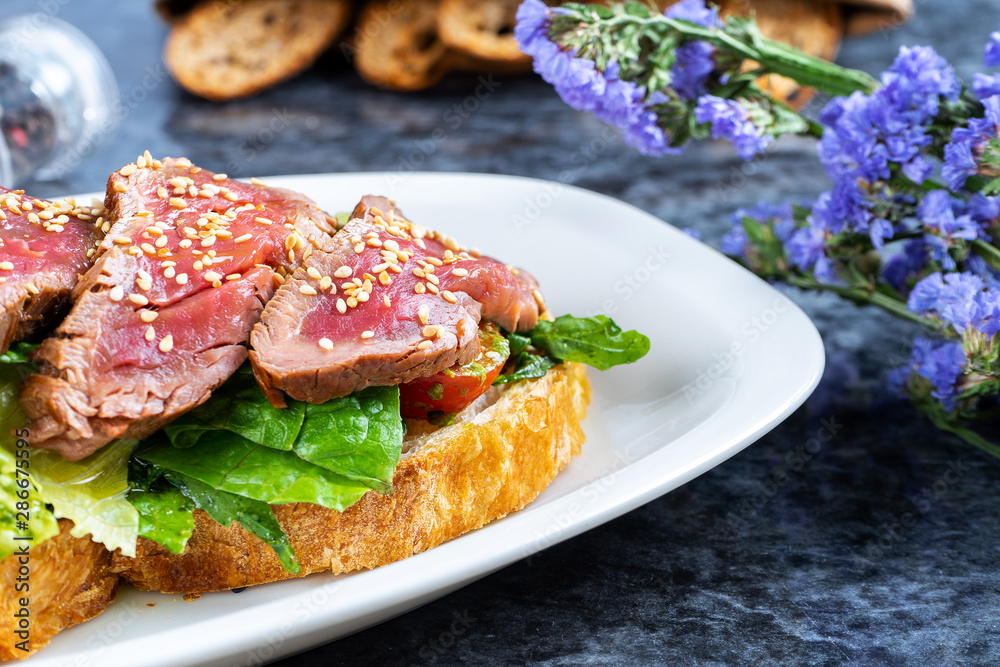 Close up view on delicious italian bruschetta with roast beef, lettuce, cherry tomato and sauce. Italian cuisine. Selective focus on sandwich with meat on marble table. Copy space