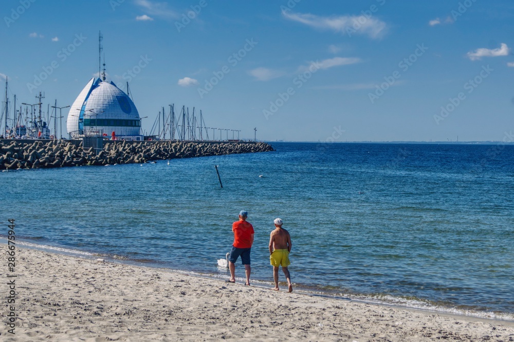 para mężczyzn spaceruje wzdłuż plaży w kierunku portu Hel, widoczne umocnienia brzegu w porcie i maszty żaglówek