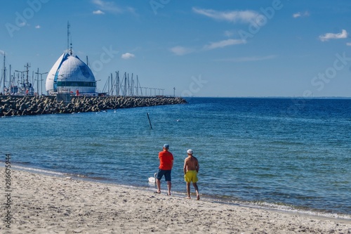 Obraz na płótnie para mężczyzn spaceruje wzdłuż plaży w kierunku portu Hel, widoczne umocnienia brzegu w porcie i maszty żaglówek w salonie