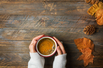 Young caucasian woman girl in knitted white sweater holds in hands mug with freshly brewed coffee with appetizing crema dry orange yellow leaves on aged wood kitchen table. Cozy fall autumn atmosphere