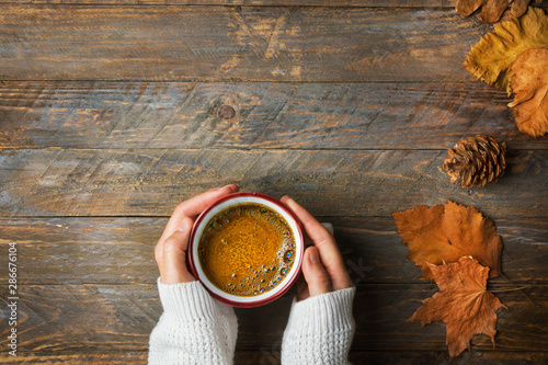 Młoda caucasian kobiety dziewczyna w trykotowym białym pulowerze trzyma w ręka kubku z świeżo parzoną kawą z apetycznej crema suchymi pomarańczowymi żółtymi liśćmi na starzejącym się drewnianym kuchennym stole. Przytulna jesienna atmosfera