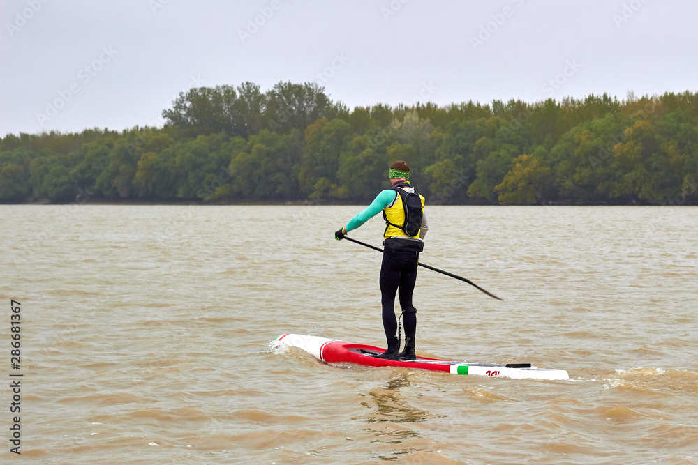 Athlete paddling SUP (Stand up paddle board) at Danube river at cold weather against overcast sky. Concept of water tourism, water sport, healthy lifestyle and recreation