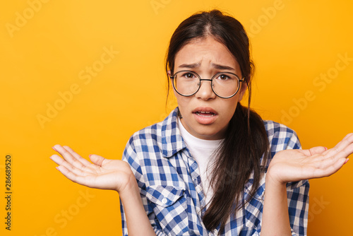 Confused teenage girl in glasses posing isolated over yellow wall background.