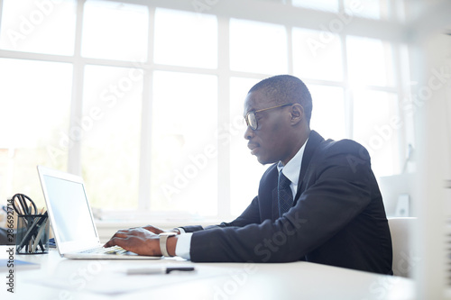 Concentrated African businessman in eyeglasses typing on laptop keyboard while sitting at his workplace at office