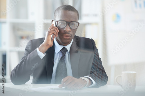 Serious African office worker in eyeglasses and in suit sitting at his workplace at office and has a phone call on mobile phone